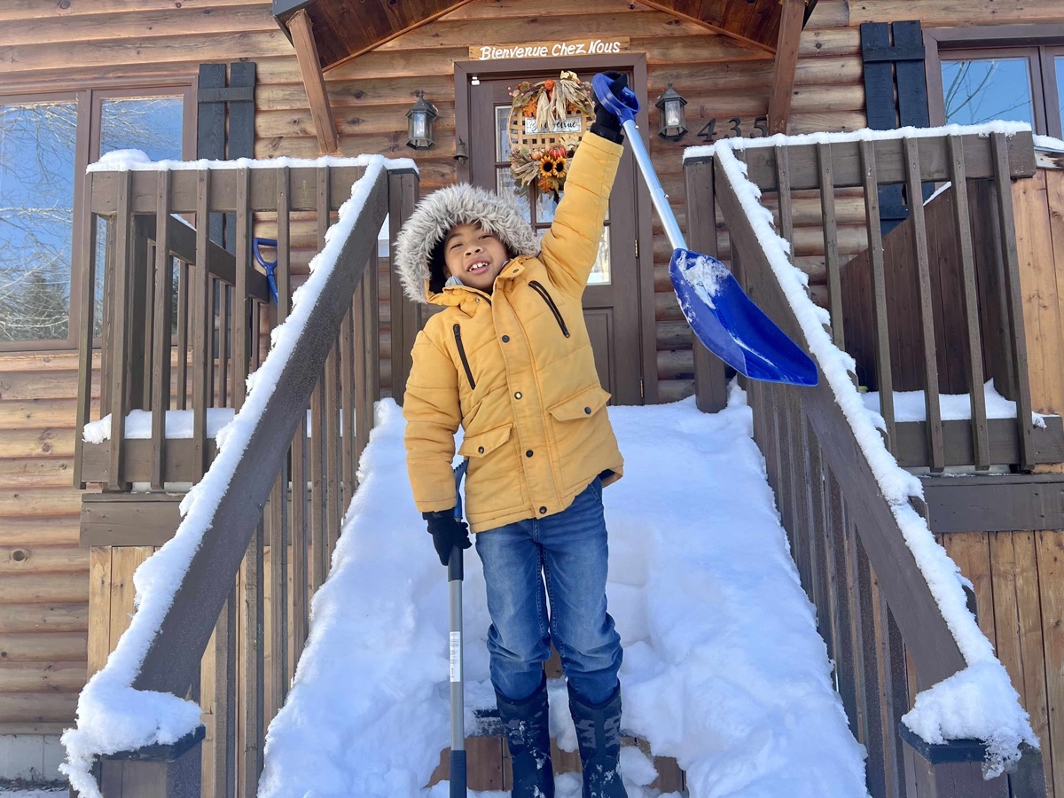A boy raising a snow shovel in triumph on the snowy steps of a log cabin